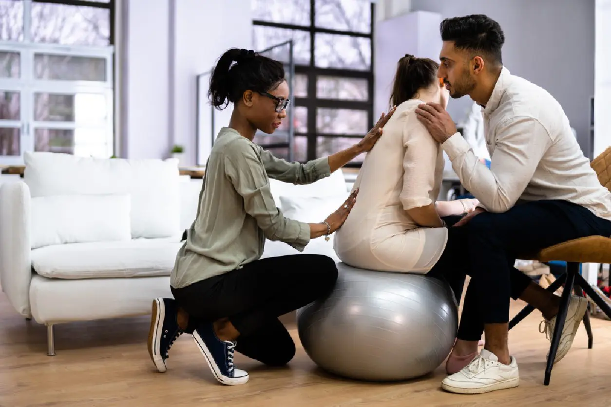 People supporting woman on exercise ball indoors.