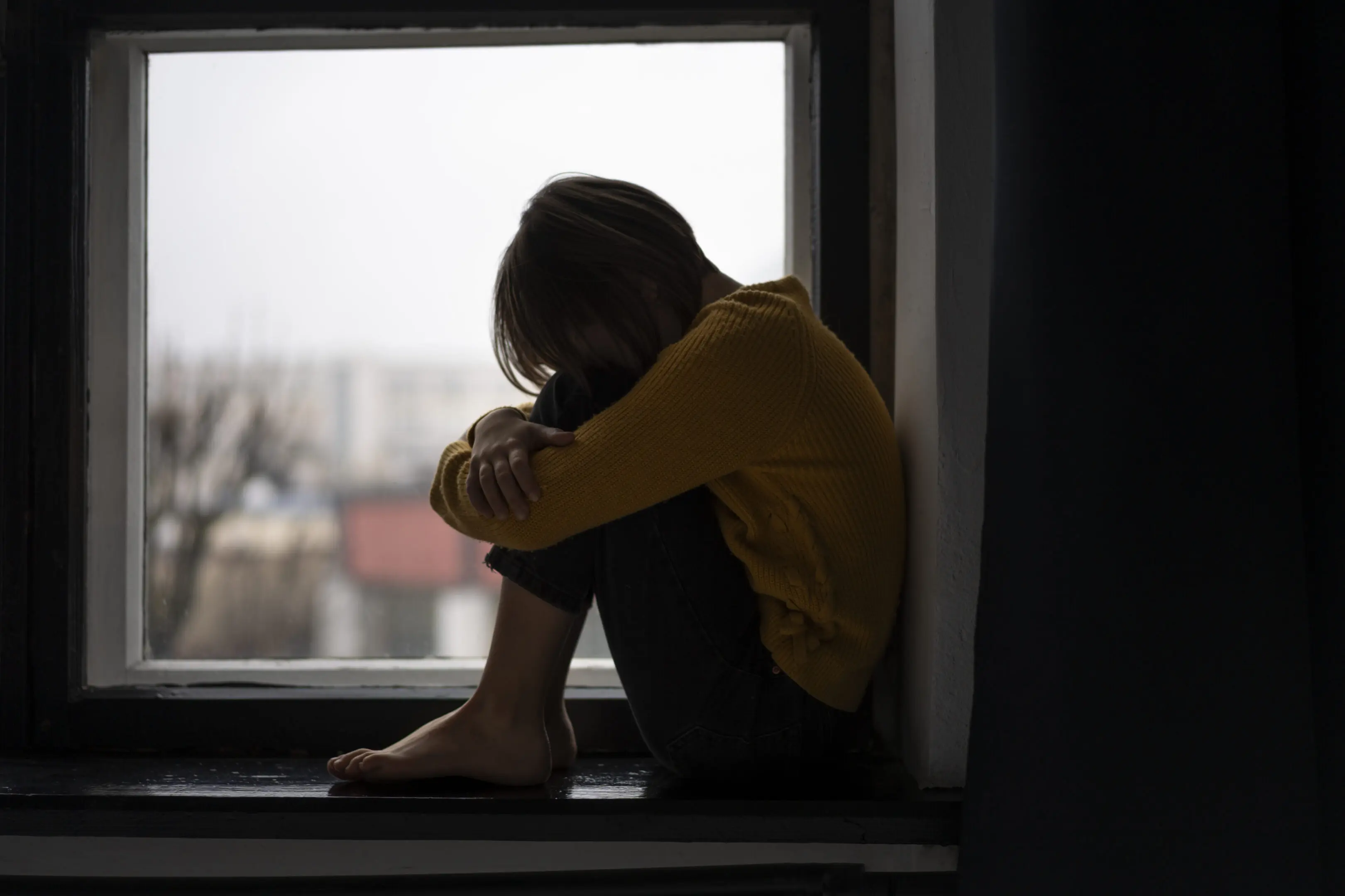 A silhouette of a person sitting by a window, hugging their knees.